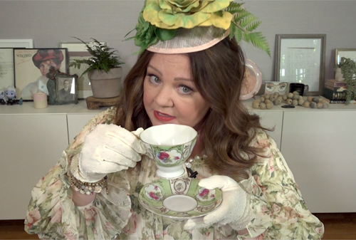 Melissa McCarthy trägt ein opulentes Kleid, einen Fascinator-Hut und weiße Handschuhe und hält eine Teetasse in der Hand