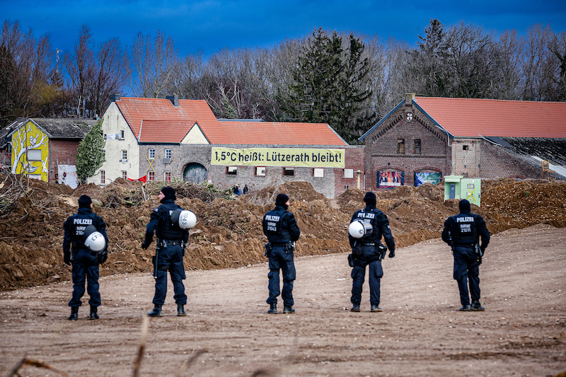 Kette aus Polizisten vor dem Dord Lützerath. Auf einem Banner an einem Haus steht: "1,5 Grad heißt: Lützerath bleibt!"