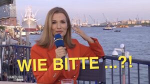 NDR-Moderatorin beim Hamburger Hafengeburtstag deutet auf ihr Ohr. Davor der Schriftzug: WIE BIETTE ...?!"