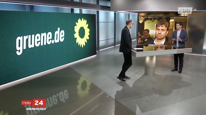 rbb-Reporter und "Abendschau"-Moderator im Studiogespräch. Hinter ihnen: das Logo der Grünen und ein Foto von Stefan Gelbhaar.