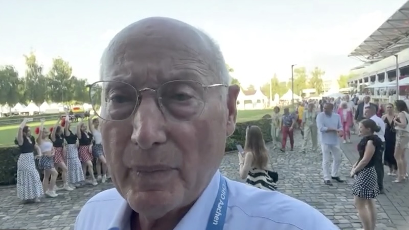 Der Journalist Stefan Aust steht am Rande des Aachener Pferdestadions und spricht in sein Smartphone. Hinter ihm posieren junge Leute fürs Foto.