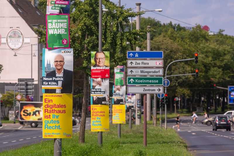 Befahrene Straße in Köln. An Masten hängen verschiedene Wahlplakate, unter anderem von SPD, CDU und FDP.