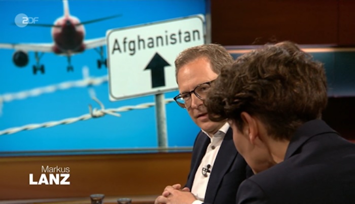 Talksrunde bei „Markus Lanz“, im Hintergrund ein Foto mit einem Schild, auf dem „Afghanistan“ steht nebst einem Pfeil, der nach oben deutet, gen Himmel, im Hintergrund ein Stück Stacheldraht und ein abhebendes Flugzeug vor strahlend blauem Himmel.