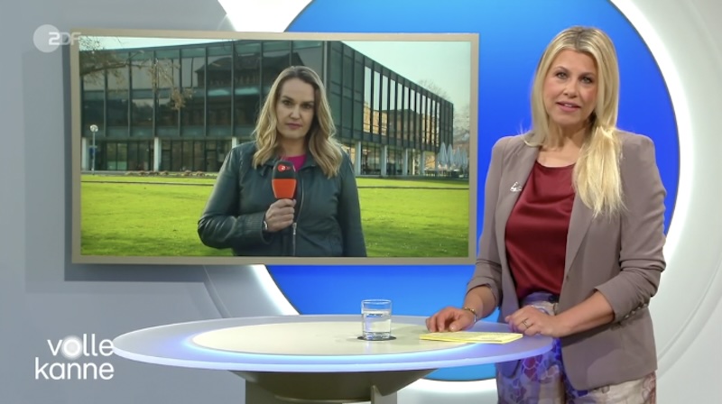 „Volle Kanne“-Moderatorin Nadine Krüger steht an einem Tisch vor einem Bildschirm, auf dem die Korrespondentin Sandra Susanka vor dem Landtag in Stuttgart steht.