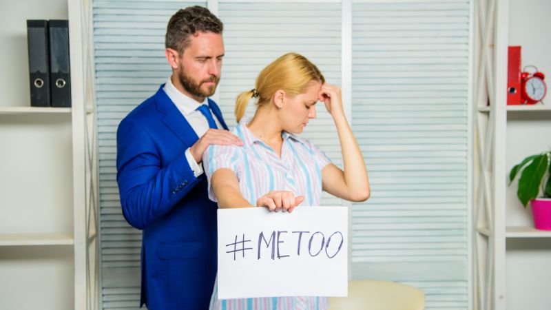 Mann im Büro legt seiner Kollegin eine Hand auf die Schulter, während sie ein Papier hochhält, auf dem „metoo“ steht