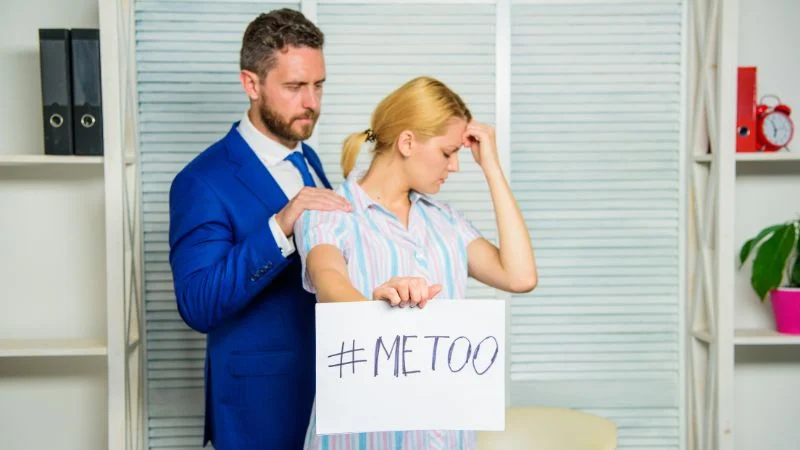 Mann im Büro legt seiner Kollegin eine Hand auf die Schulter, während sie ein Papier hochhält, auf dem „metoo“ steht