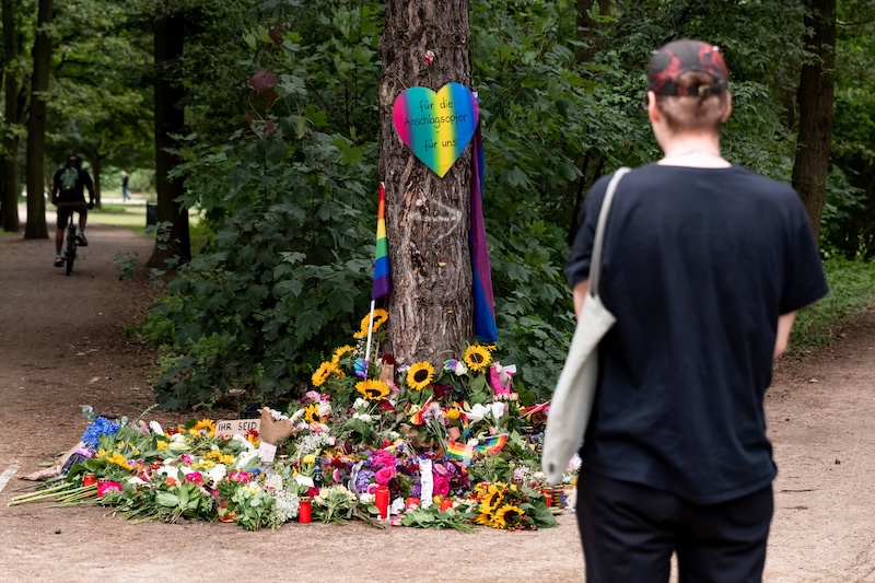 Eine Person steht vor einer Gedenkstelle mit Blumen und Regenbogenflaggen im Berliner Tiergarten. Auf einem Schild steht: „Für die Anschlagsopfer – für uns“.