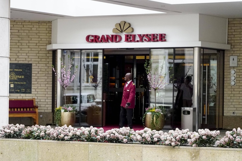 Gläserner Eingang des Hotels Grand Elysée in Hamburg. Im Vordergrund: eine Reihe mit Blumen in Blumenkästen.