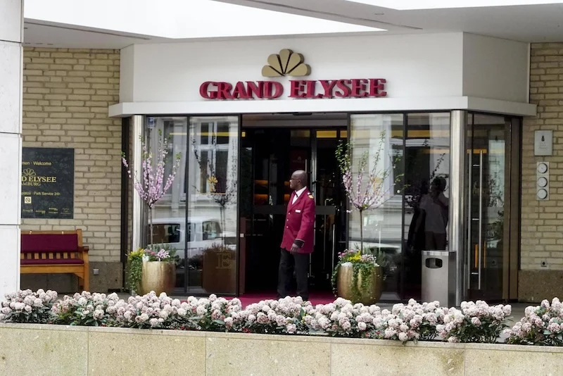 Gläserner Eingang des Hotels Grand Elysée in Hamburg. Im Vordergrund: eine Reihe mit Blumen in Blumenkästen.
