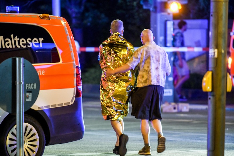 Von hinten zu sehen: Eine Person legt den Arm um eine andere, die in eine goldene Rettungsdecke gehüllt ist. Daneben steht ein Krankenwagen.