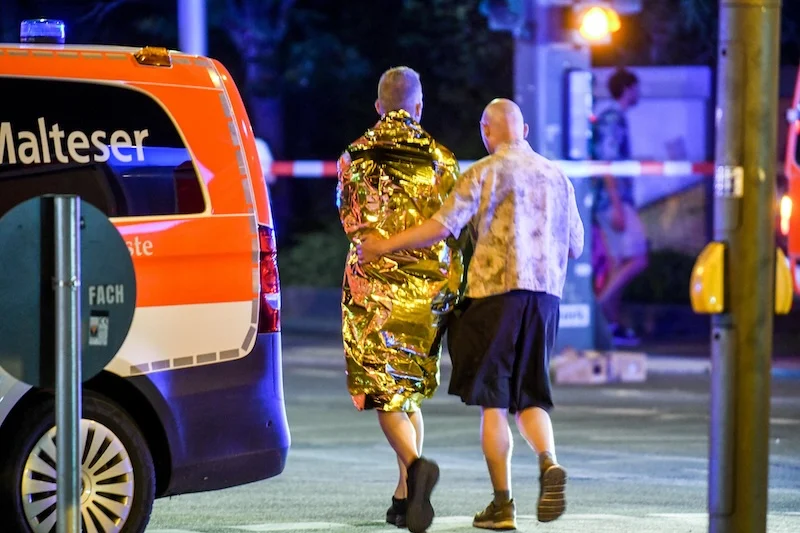 Von hinten zu sehen: Eine Person legt den Arm um eine andere, die in eine goldene Rettungsdecke gehüllt ist. Daneben steht ein Krankenwagen.
