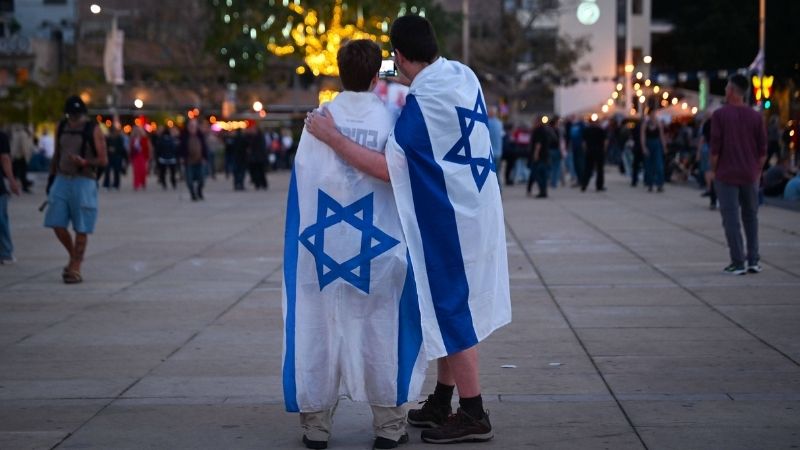Zwei junge Männer auf einem Platz in Tel Aviv machen ein Selfie mit zwei israelischen Flaggen