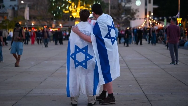 Zwei junge Männer auf einem Platz in Tel Aviv machen ein Selfie mit zwei israelischen Flaggen
