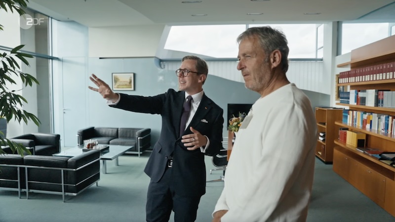 Philipp Amthor steht in seinem Büro und deutet mit der Hand in die Ferne. Neben ihm steht der Mann, der den Strandkorb geliefert hat. Im Hintergrund zu sehen: eine Sitzgruppe aus schwarzem Leder.
