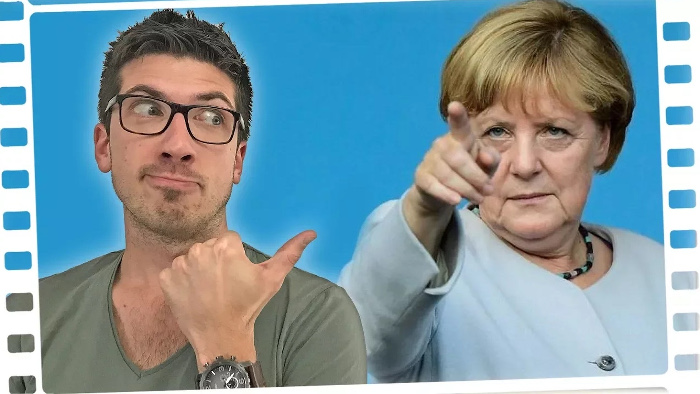 Ein Mann mit kurzen Haaren und Brille (links) deutet auf Angela Merkel, die mit dem Zeigefinger nach vorne zeigt.
