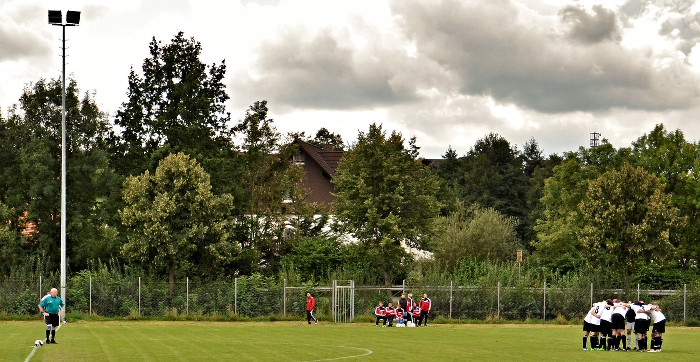 Totale eines Fußballplatzes, links steht der Schiedsrichter in der Spielfeldmitte, rechts die Mannschaft im Kreis.