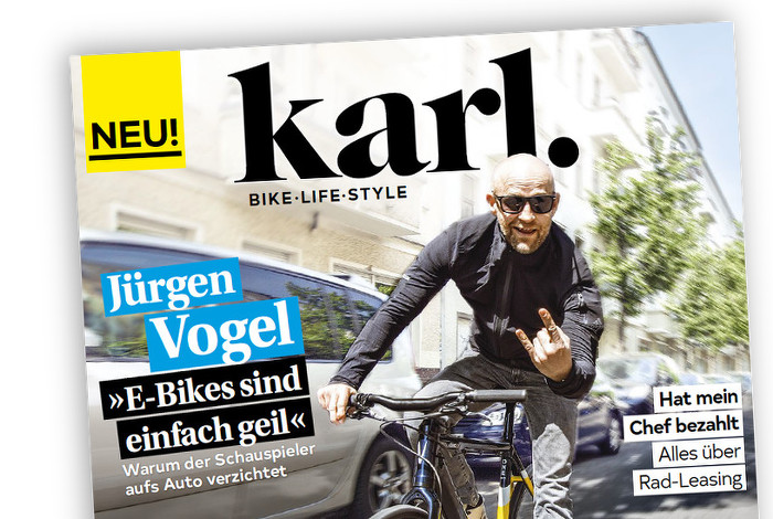 Jürgen Vogel (mit Glatze und Sonnenbrille) fährt Fahrrad und zeigt dabei Teufelshörner mit der Hand.
