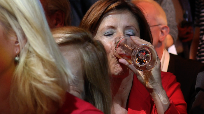 Frau mit dunklen Haaren und rotem Blazer trinkt ein Bier und hat dabei die Augen geschlossen.