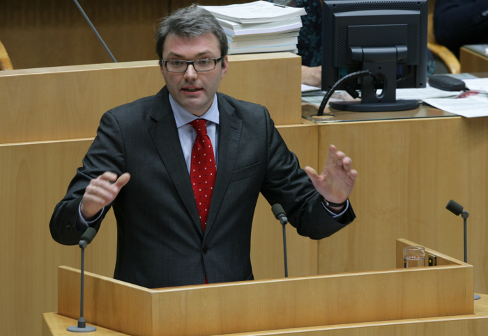 Mann mit Brille, Anzug und roter Krawatte hält eine Rede an einem Pult.