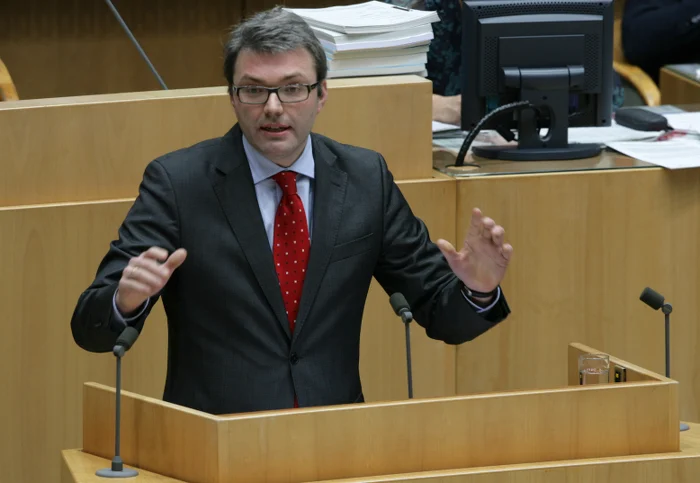 Mann mit Brille, Anzug und roter Krawatte hält eine Rede an einem Pult.