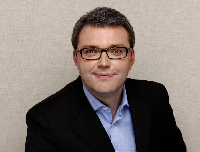 Mann mit Brille, dunklen Haaren, Sakko, Hemd schaut in die Kamera.