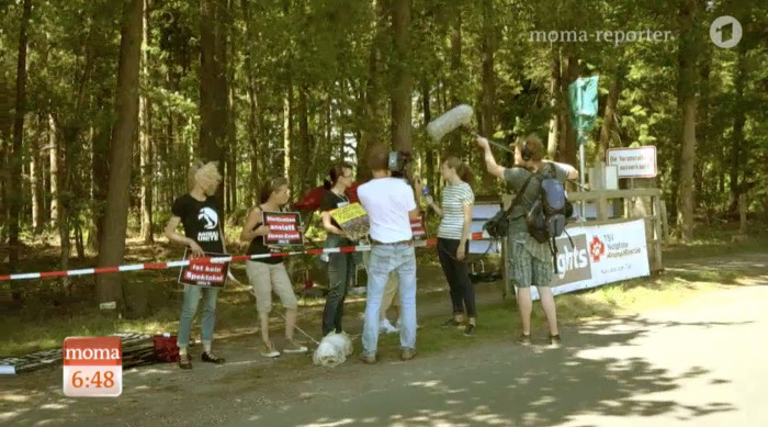 Reporterin und Kamerateam stehen vor vier Demonstranten mit Schildern und interviewen sie.