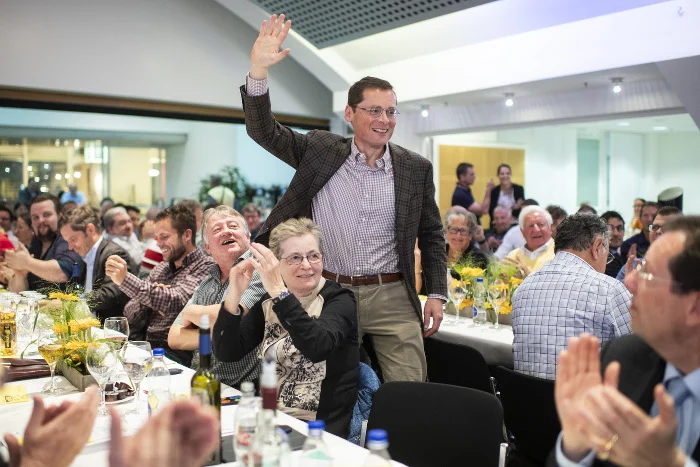 Mann mit Brille und im Anzug geht winkend durch Tischreihen in einem Saal.
