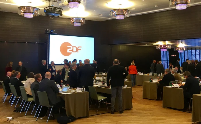 Großer Saal mit Tischen, an denen Menschen sitzen oder stehen. Im Hintergrund eine Leinwand mit ZDF-Logo.